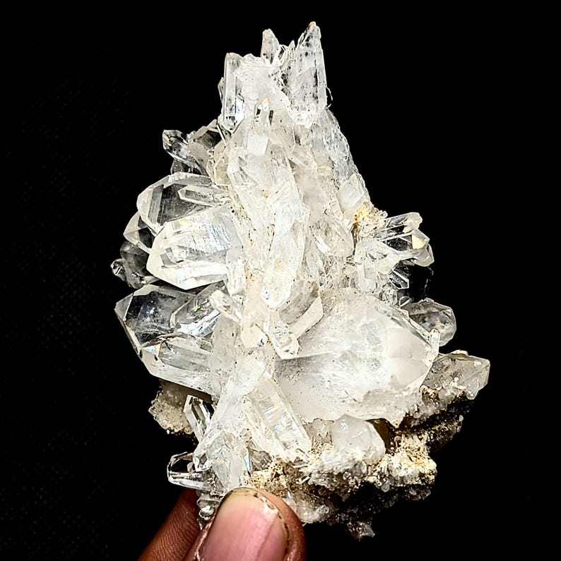 May include: A cluster of clear, translucent quartz crystals with sharp, pointed terminations growing from a rough, greyish-brown mineral matrix. The crystalline structure features multiple facets that reflect light, creating a bright, jagged appearance.
