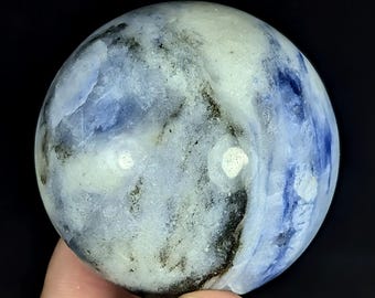 May include: A polished spherical stone featuring a marbled pattern of white, translucent light blue, and deep navy blue sections with dark grey and black mineral inclusions throughout the surface, creating a natural and varied textured appearance.