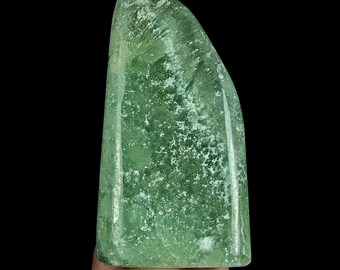 May include: A polished, semi-translucent green mineral specimen with a smooth, rounded top and a flat base. The stone features internal white crystalline inclusions and a mottled texture throughout, displaying varying shades of light and dark green.