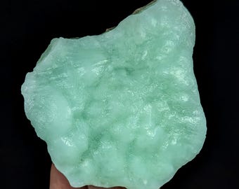 May include: A raw, translucent mint green mineral specimen with a botryoidal, bumpy surface texture. The stone features irregular edges and a crystalline, semi-glossy appearance that catches the light, showcasing varying shades of pale green.