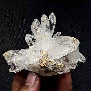 May include: A cluster of clear, translucent quartz crystals featuring sharp, pointed terminations and a jagged, multifaceted structure. The base of the mineral specimen displays earthy, tan-colored inclusions and rough, textured rock matrix.