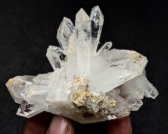 May include: A cluster of clear, translucent quartz crystals featuring sharp, pointed terminations and a jagged, multifaceted structure. The base of the mineral specimen displays earthy, tan-colored inclusions and rough, textured rock matrix.