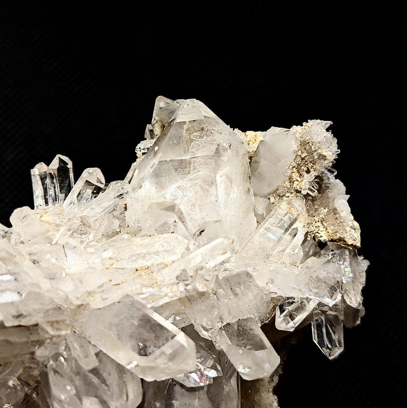 May include: A cluster of clear, translucent quartz crystals featuring sharp, pointed terminations and multifaceted surfaces. The mineral formation displays a mix of large and small prismatic structures attached to a rough, light-colored rocky matrix.