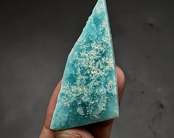 May include: A triangular-shaped piece of raw turquoise-colored mineral with a rough, textured surface. The stone features a vibrant blue-green hue with natural white and cream-colored inclusions scattered across its uneven, crystalline face.