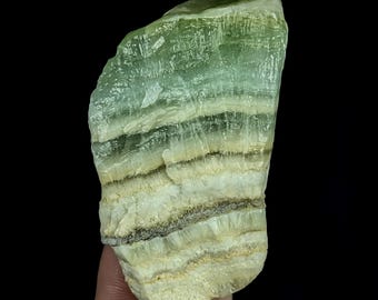 May include: A raw, unpolished mineral specimen featuring horizontal bands of pale green, cream, and dark grey. The stone has a translucent, fibrous texture with a layered appearance, showing natural variations in color and opacity throughout its surface.