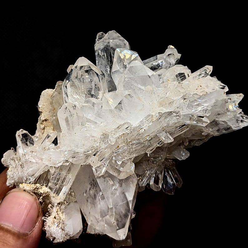 May include: A cluster of clear, translucent quartz crystals with sharp, pointed terminations growing from a rough, light-colored mineral matrix. The crystalline structure features multiple facets and varying sizes, creating a jagged, natural formation.