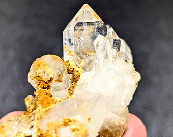 May include: A cluster of clear, pointed quartz crystals featuring sharp, transparent terminations. The mineral formation displays natural inclusions and patches of earthy, golden-brown iron oxide staining concentrated along the base and sides.