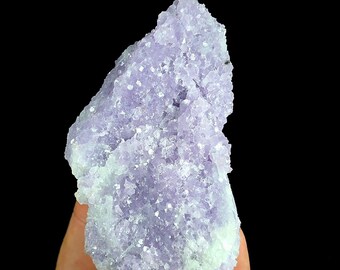 May include: A raw, jagged mineral specimen featuring a cluster of light purple, translucent crystals. The formation has a rough, uneven texture with sparkling facets throughout, transitioning into a pale, whitish base at the lower edges.