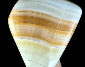 May include: A polished, smooth stone specimen featuring horizontal bands of translucent white, cream, and warm amber-brown colors. The mineral has a rounded, irregular shape with a glossy, reflective surface that highlights the natural layered patterns.