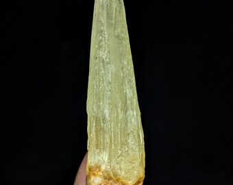 May include: A translucent, pale yellow, elongated mineral crystal with a vertical, striated texture. The specimen features a tapered, pointed top and a rough, brownish-orange base, showcasing natural crystalline formations and light refraction.