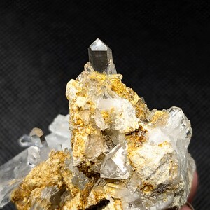 May include: A cluster of clear, pointed quartz crystals emerging from a textured, golden-brown mineral matrix. The central crystal stands vertically, surrounded by smaller, jagged crystalline formations with translucent edges and earthy deposits.