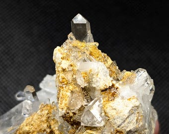 May include: A cluster of clear, pointed quartz crystals emerging from a textured, golden-brown mineral matrix. The central crystal stands vertically, surrounded by smaller, jagged crystalline formations with translucent edges and earthy deposits.