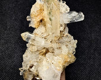 May include: A cluster of clear and translucent quartz crystals featuring a prominent central point with a subtle green inclusion. The mineral formation displays jagged, multi-directional points with golden-brown earthy deposits nestled between the crystalline structures.