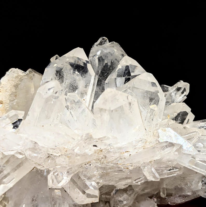 May include: A cluster of clear, translucent quartz crystals featuring sharp, pointed terminations and multifaceted surfaces. The mineral formation displays a natural, jagged structure with varying sizes of prismatic points extending outward from a central base.