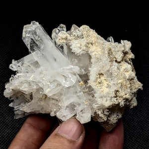 May include: A cluster of clear, translucent quartz crystals featuring sharp, pointed terminations. One side of the mineral specimen displays a rough, textured matrix with earthy, beige-colored mineral deposits attached to the crystalline structure.
