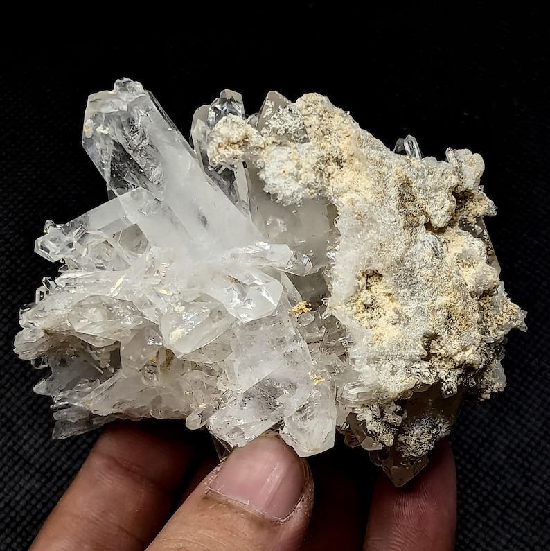 May include: A cluster of clear, translucent quartz crystals featuring sharp, pointed terminations. One side of the mineral specimen displays a rough, textured matrix with earthy, beige-colored mineral deposits attached to the crystalline structure.