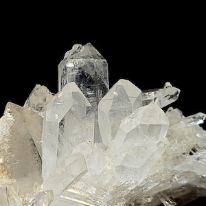 Può includere: Un gruppo di cristalli di quarzo trasparenti e traslucidi con molteplici terminazioni appuntite e superfici sfaccettate. La formazione minerale mostra prismi esagonali di varie dimensioni che crescono da una base comune.