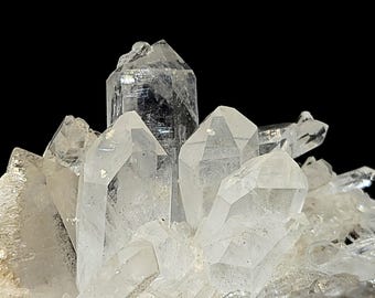 May include: A cluster of clear, translucent quartz crystals featuring multiple pointed terminations and faceted surfaces. The mineral formation displays varying sizes of hexagonal prisms growing from a common base, with visible internal inclusions.