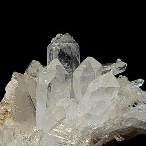 May include: A cluster of clear, translucent quartz crystals featuring multiple pointed terminations and faceted surfaces. The mineral formation displays varying sizes of hexagonal prisms growing from a common base against a black background.