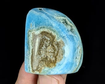 May include: A polished, semi-circular blue hemimorphite stone featuring a prominent, textured brown and tan mineral inclusion in the center. The stone displays vibrant shades of sky blue with translucent crystalline edges and a smooth surface.