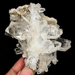 May include: A cluster of clear, translucent quartz crystals with sharp, pointed terminations and multifaceted surfaces. The mineral specimen features a rough, light-colored matrix at the top, with individual crystalline structures radiating outward.