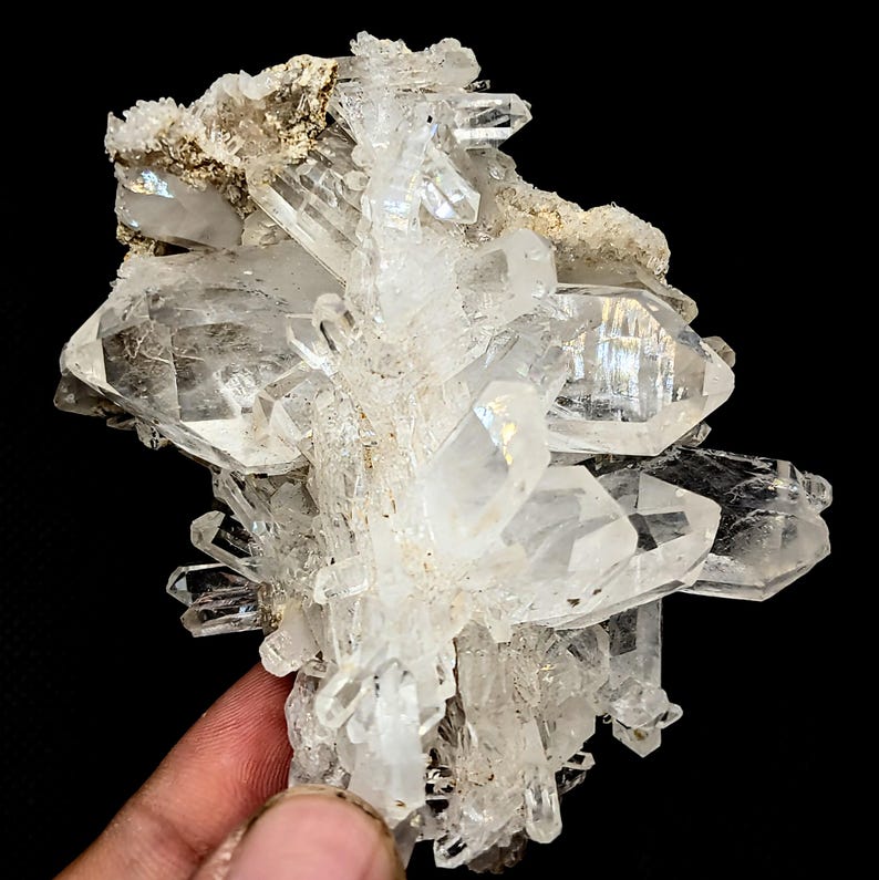 May include: A cluster of clear, translucent quartz crystals with sharp, pointed terminations and multifaceted surfaces. The mineral specimen features a rough, light-colored matrix at the top, with individual crystalline structures radiating outward.