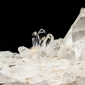 May include: A cluster of clear, translucent quartz crystals featuring sharp, pointed terminations and multifaceted surfaces. The mineral formation displays varying sizes of crystalline structures with a bright, reflective quality against a dark background.