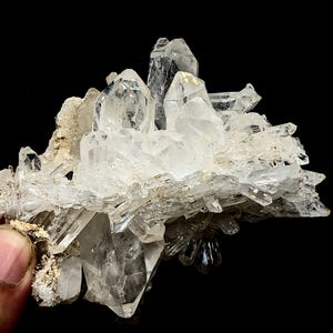 May include: A cluster of clear, pointed quartz crystals growing from a rough, tan-colored mineral matrix. The specimen features multiple sharp, translucent points of varying sizes that radiate outward, showcasing natural geometric facets.