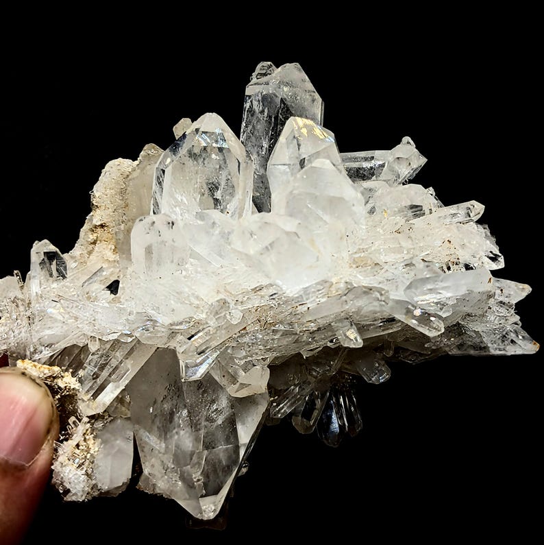 May include: A cluster of clear, pointed quartz crystals growing from a rough, tan-colored mineral matrix. The specimen features multiple sharp, translucent points of varying sizes that radiate outward, showcasing natural geometric facets.