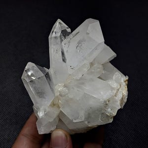 May include: A cluster of clear, translucent quartz crystals with sharp, pointed terminations. The mineral specimen features multiple hexagonal prisms growing together from a common base, displaying natural facets and internal inclusions.