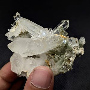 May include: A cluster of clear, translucent quartz crystals with sharp, pointed terminations. The mineral specimen features a mix of large, prismatic crystals and smaller, jagged formations with earthy, greenish-grey mineral inclusions.
