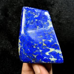May include: A polished piece of lapis lazuli stone featuring a deep royal blue base color with prominent white calcite veining and scattered golden pyrite inclusions. The stone has a smooth, reflective surface and an irregular, angular shape.