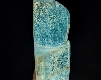 May include: A polished slab of blue larimar stone featuring intricate, cloud-like patterns in shades of sky blue, turquoise, and white, with subtle tan mineral inclusions visible across the smooth, semi-translucent surface of the mineral specimen.