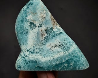 May include: A polished slab of blue larimar stone featuring a triangular shape with rounded edges. The surface displays a complex pattern of swirling turquoise and white mineral inclusions, creating a textured appearance with varying shades of blue.