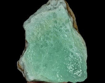 May include: A raw, unpolished mineral specimen featuring a translucent, mint-green crystalline surface with a textured, bumpy appearance. The stone has irregular, jagged edges with hints of brown and white mineral deposits along the outer perimeter.