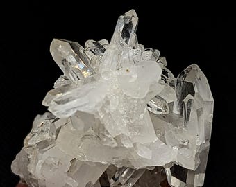 May include: A cluster of clear, translucent quartz crystals with sharp, pointed terminations and multifaceted surfaces. The mineral formation features a central grouping of smaller, opaque white crystals surrounded by larger, transparent points.