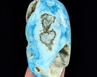 May include: An oval-shaped polished Larimar stone featuring a vibrant sky-blue color with white marbling and a central, textured, brownish-grey inclusion. The surface displays natural indentations and a smooth, glossy finish on the blue areas.