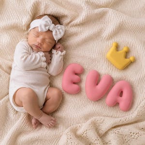 May include: A sleeping infant wearing a white ribbed long-sleeve bodysuit and a white headband with a bow. Beside the infant are three pink, rounded, bubble-style letters spelling EVA and a small, yellow, three-pointed crown-shaped object.