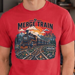 Può includere: Una t-shirt rossa a maniche corte con un'illustrazione grafica di una locomotiva a vapore sui binari. Il testo MERGE TRAIN è stampato sopra il treno e un cartello con la scritta NEXT STOP PRODUCTION è visibile vicino ai binari sotto un cielo al tramonto.