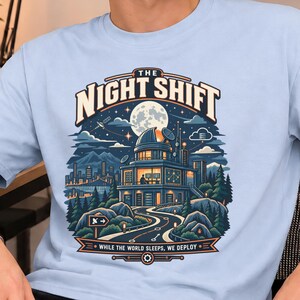 Op de afbeelding: Een lichtblauw t-shirt met een grafische illustratie van een observatorium onder een volle maan en sterrenhemel. De tekst luidt THE NIGHT SHIFT bovenaan en WHILE THE WORLD SLEEPS, WE DEPLOY onderaan, met een kronkelende weg naar het gebouw.