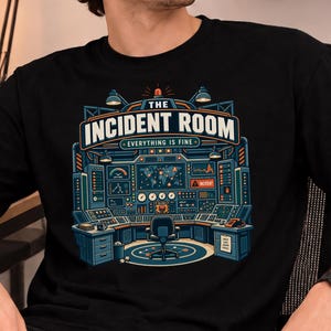 May include: A black t-shirt featuring a graphic print of a control room console with monitors, dials, and a central chair. The text reads THE INCIDENT ROOM and EVERYTHING IS FINE. The design uses blue, orange, and white colours on a black background.