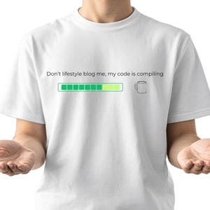 May include: A white short-sleeved t-shirt featuring the black text "Don't lifestyle blog me, my code is compiling" above a green progress bar and a small line-art illustration of a coffee mug.