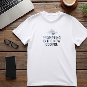 Pode incluir: Uma t-shirt branca de manga curta com um gráfico de um cérebro humano com linhas de circuitos digitais e o texto "PROMPTING IS THE NEW CODING." impresso em letras maiúsculas azul-escuras no centro do peito da peça de vestuário.