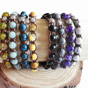 Op de afbeelding: Een collectie van acht kralenarmbanden met ronde stenen in diverse kleuren, waaronder geel, lichtblauw, donkerblauw, zwart, grijs, paars en wit. Elke armband is gemaakt met bruin koord en bevat zilverkleurige metalen accenten.