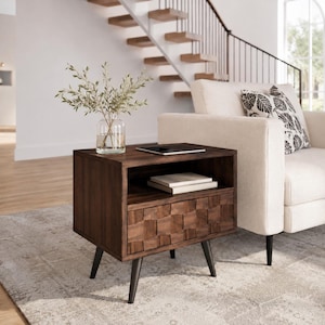 May include: A dark brown wooden nightstand with four tapered black legs, an open storage shelf containing two stacked books, and a single drawer featuring a textured, woven-style square pattern on the front face, topped with a clear glass vase.