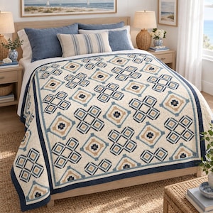 Op de afbeelding: Een crèmekleurige quilt met een herhalend geometrisch patroon in marineblauw en beige. De quilt heeft een effen marineblauwe rand en ligt over een bed met blauwe en gestreepte kussens, tussen twee nachtkastjes met lampen in geplaatst.