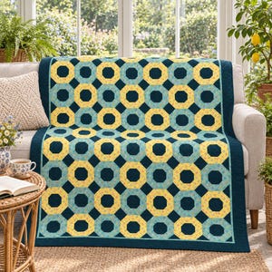Op de afbeelding: Een rechthoekige quilt met een herhalend geometrisch patroon van achthoeken in tinten geel, lichtblauw en donker marineblauw. De stof heeft een kleine bloemenprint in de vormen, afgewerkt met een effen donkerblauwe rand.