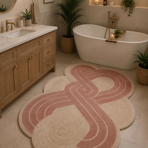 May include: An oval-shaped area rug featuring a continuous, interconnected loop pattern in soft pink and beige. The rug has a textured, tufted surface with curved lines that create a flowing, abstract design across the entire floor covering.