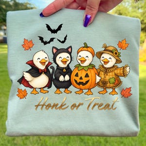 Pode incluir: Uma t-shirt verde-clara com um gráfico de quatro patos em trajes de Halloween: um vampiro, um gato preto, uma abóbora e um espantalho. Três morcegos pretos voam acima e folhas de ácer cor de laranja rodeiam as figuras.
