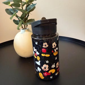May include: A black plastic water bottle featuring a repeating pattern of Mickey Mouse in various poses, including his signature red shorts and yellow shoes. The bottle has a solid black screw-top lid with a flip-up spout and a smooth cylindrical body.